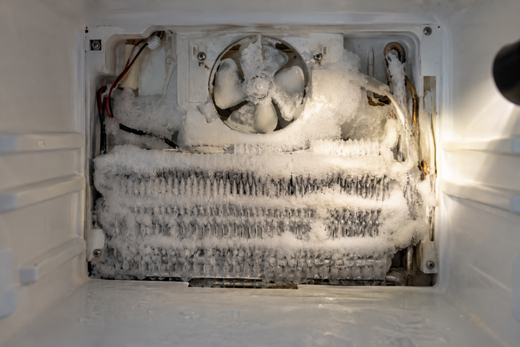 A freezer compartment with its rear panel cover removed, the evaporator coil furred with thick frost and a small fan whose blades are partly encased in ice, a flashlight beam from the side, no hands in frame.