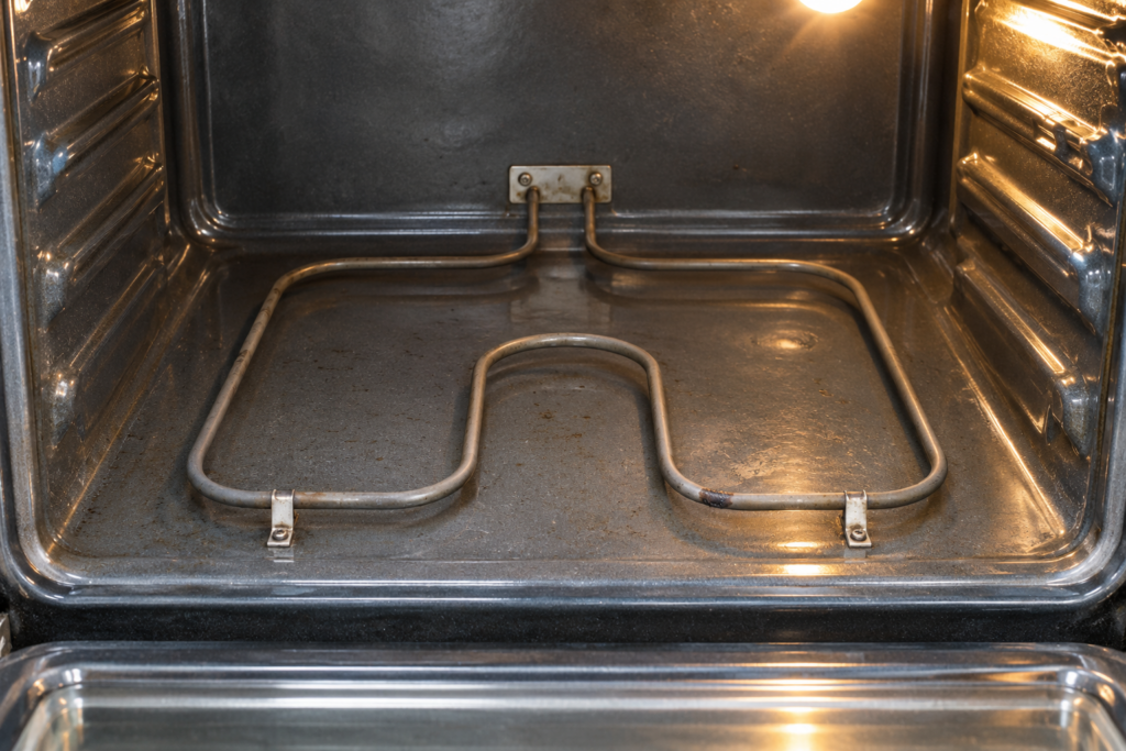 A bake element tube along the floor of an empty electric oven cavity with a small dark blistered spot on the tube, seen from above and in front, bright interior light, no hands in the frame.